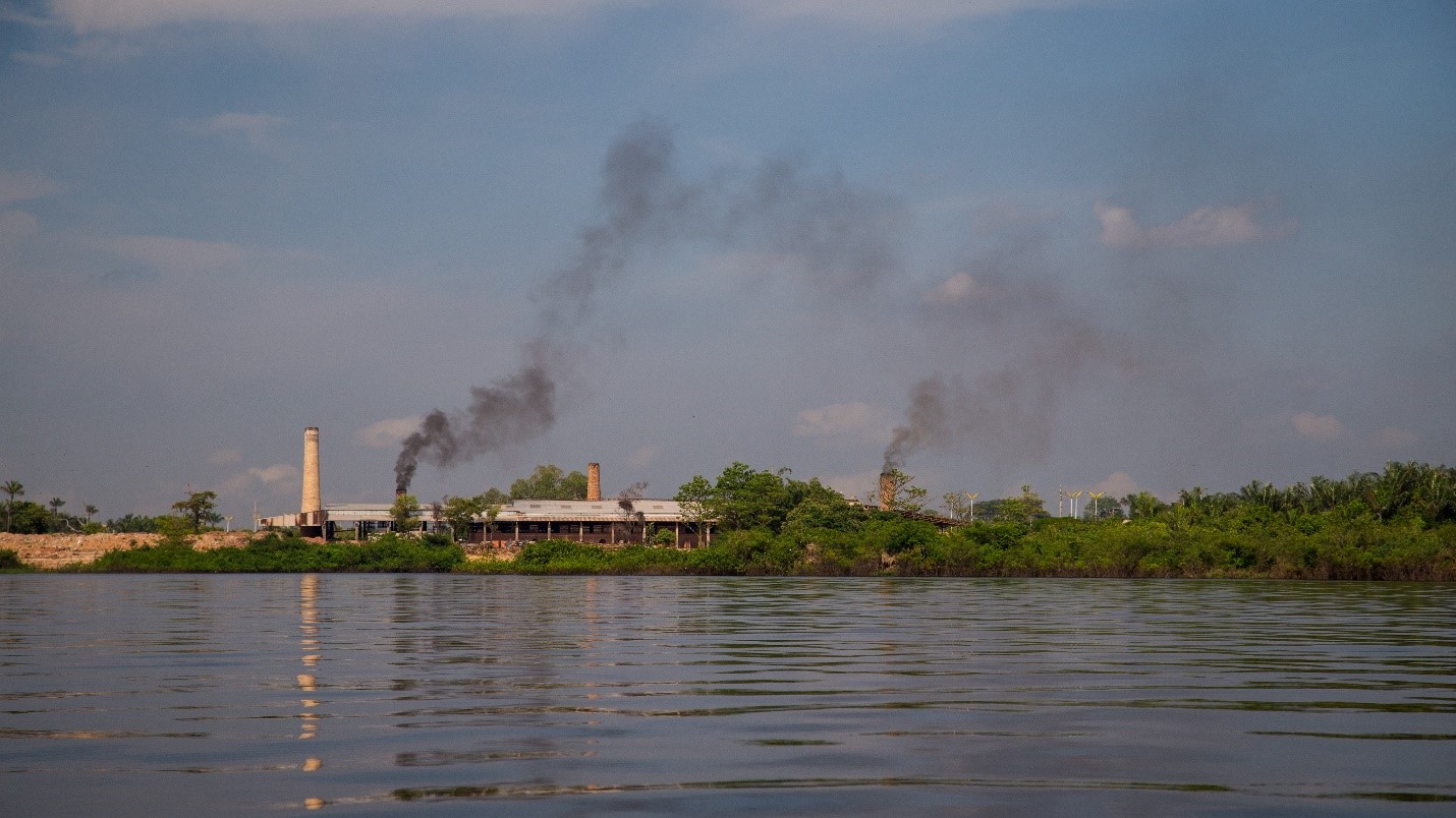 Peering out above the Amazon river waterline, several smoke stacks rise above the horizon, two of which release dark puffs of smoke.