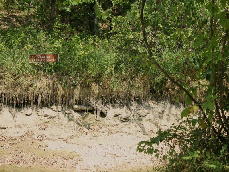 Photograph of a dried out river bed with a sign that says "caution shallow water" visible