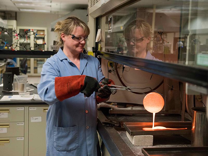 A female researcher wearing a blue lab coat and heat-resistant safety gloves pours molten glass out of a metal crucible onto a metal tray.