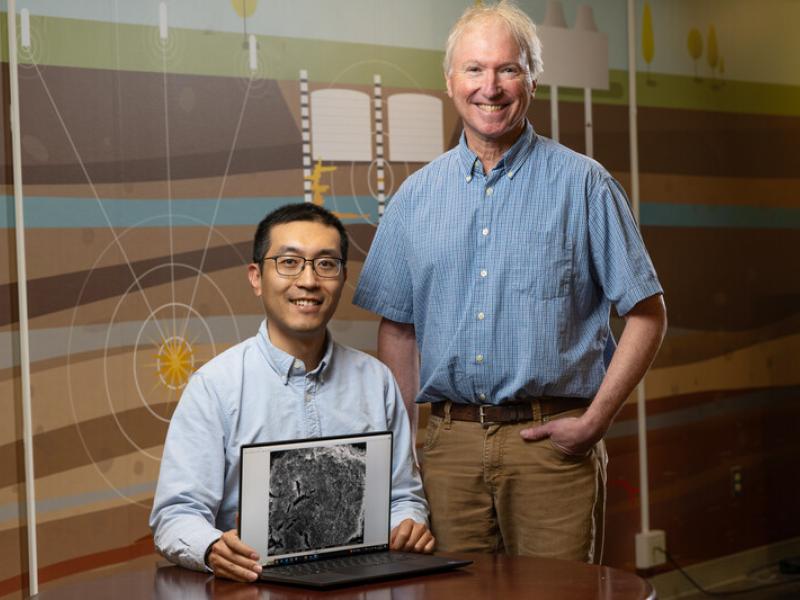 Mechanical Engineer Xiaoliang “Bryan” He holds computer with SEM image and sits while Earth Scientist Mark Rockhold stands next to him